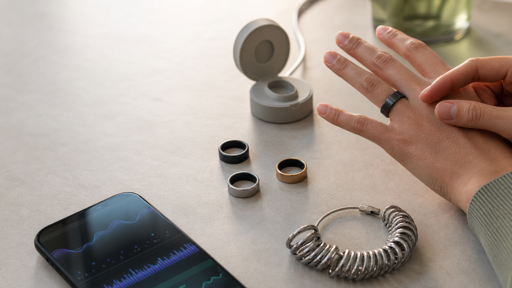 Person trying on a black smart ring beside three rings, an open charging case, a sizing gauge, and a phone showing wellness charts