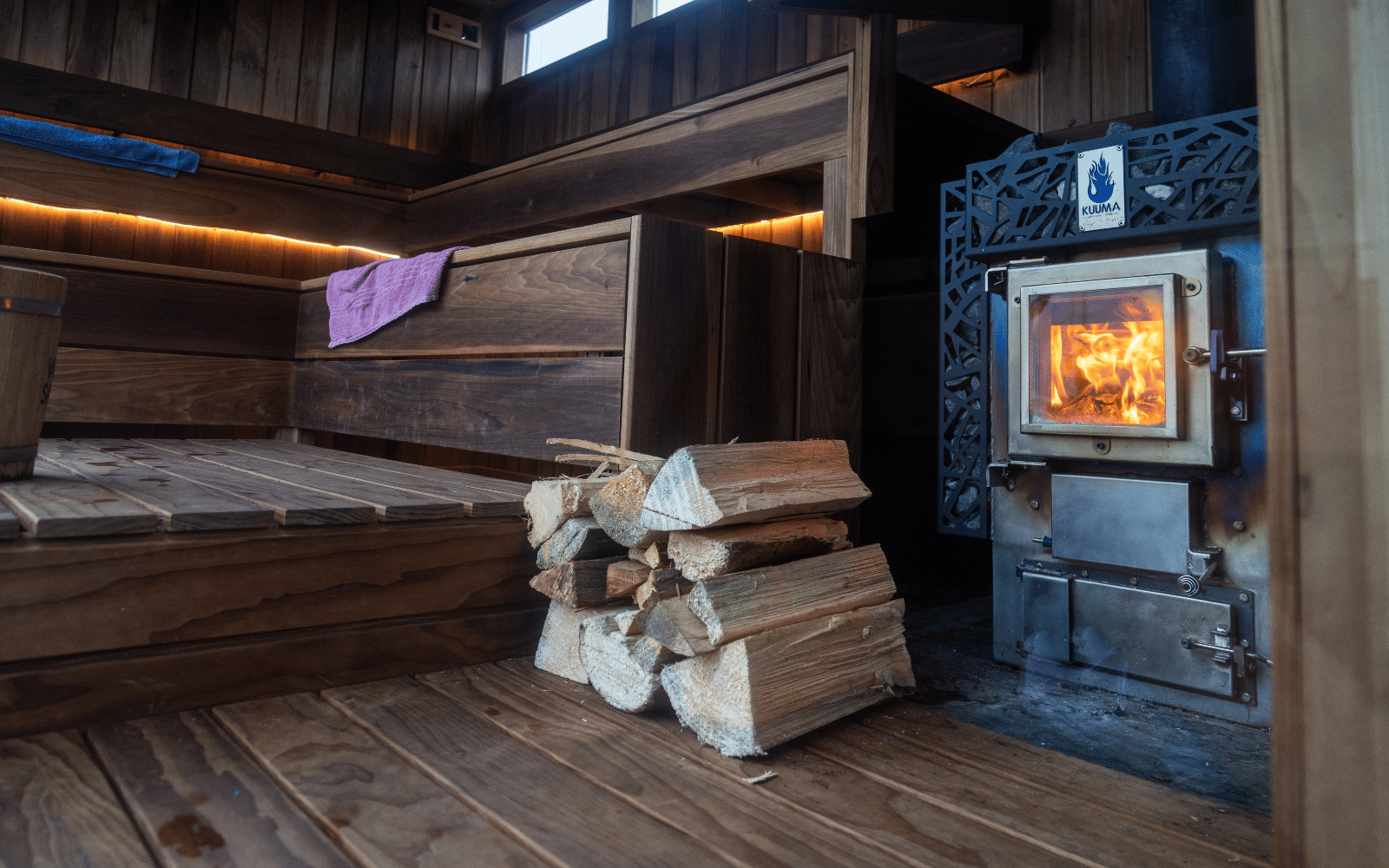 A stack of firewood in front of wooden benches and a sauna stove.
