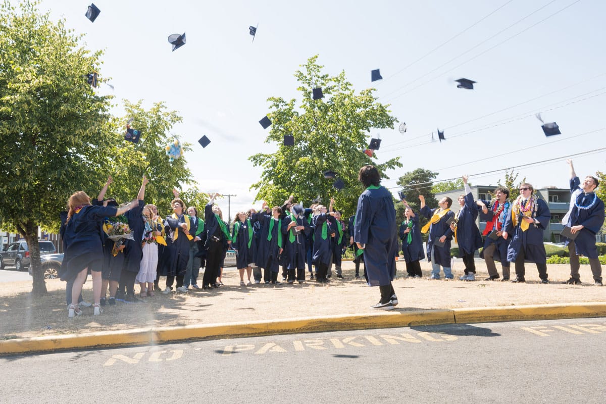 A group of students in gowns throw their caps in the air in a parking lot. 