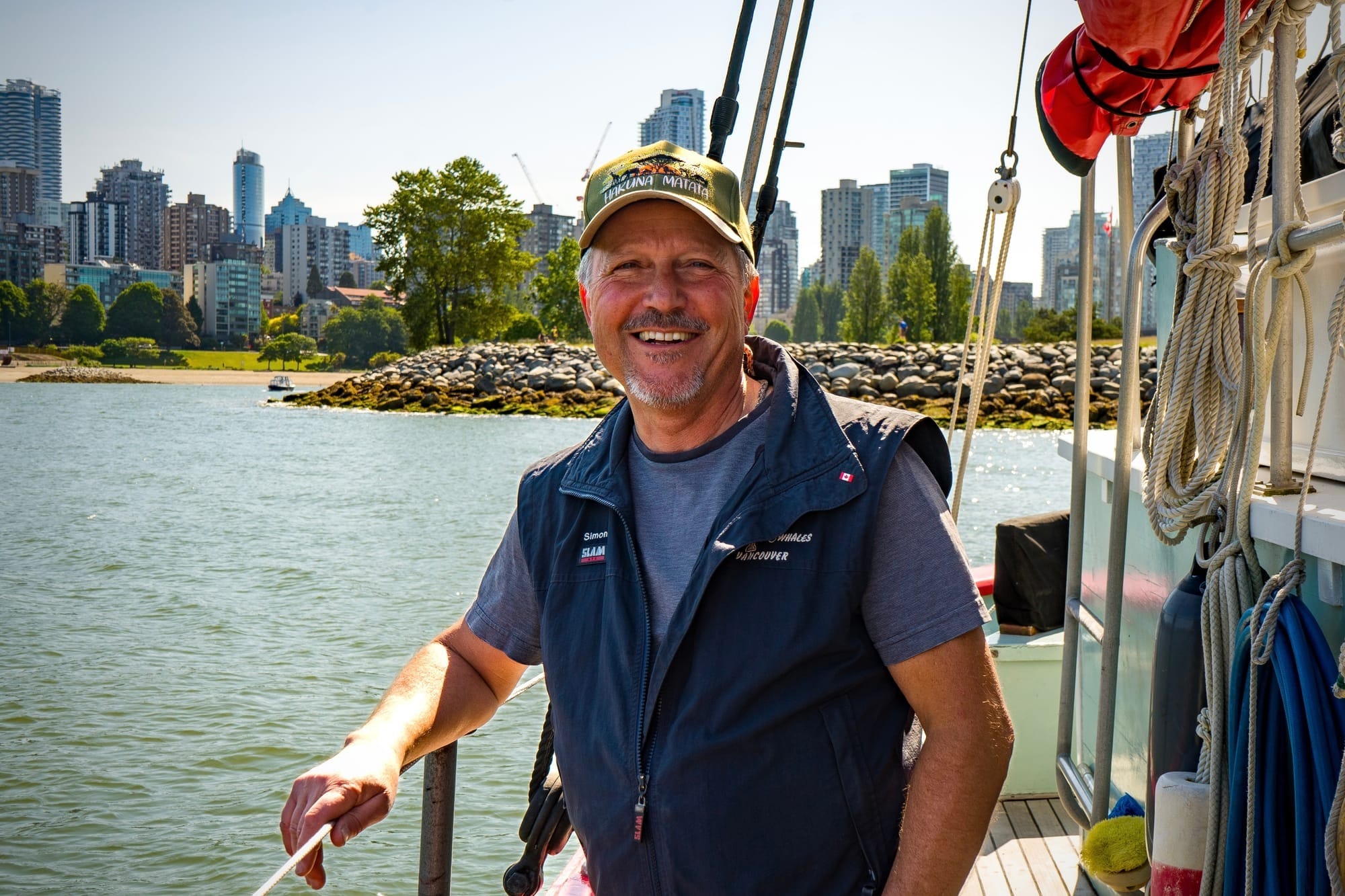 A man in a baseball cap and vest smiles on the deck of a boat with skyscrapers behind.