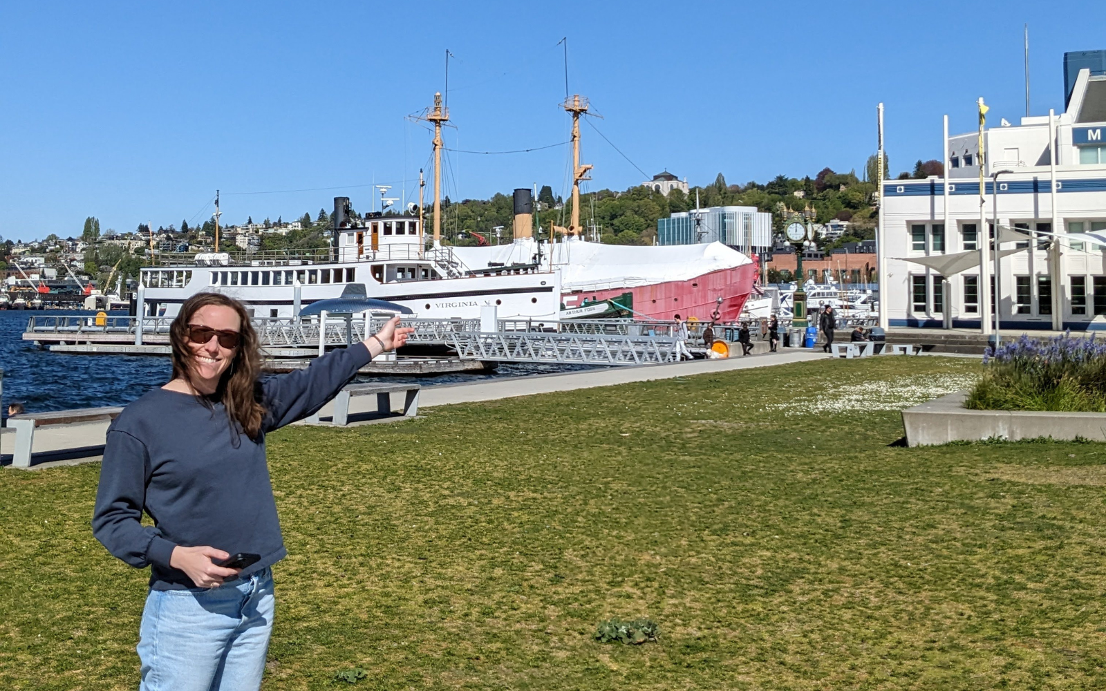 A woman wearing sunglasses gestures towards a lawn with ships and a building in the backgorund.