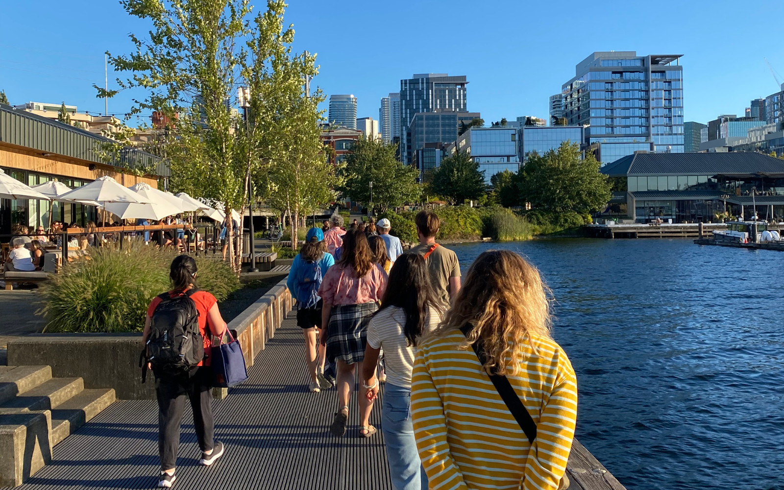 A group of people walk away on a pier with water to their right and a city skyline behind.