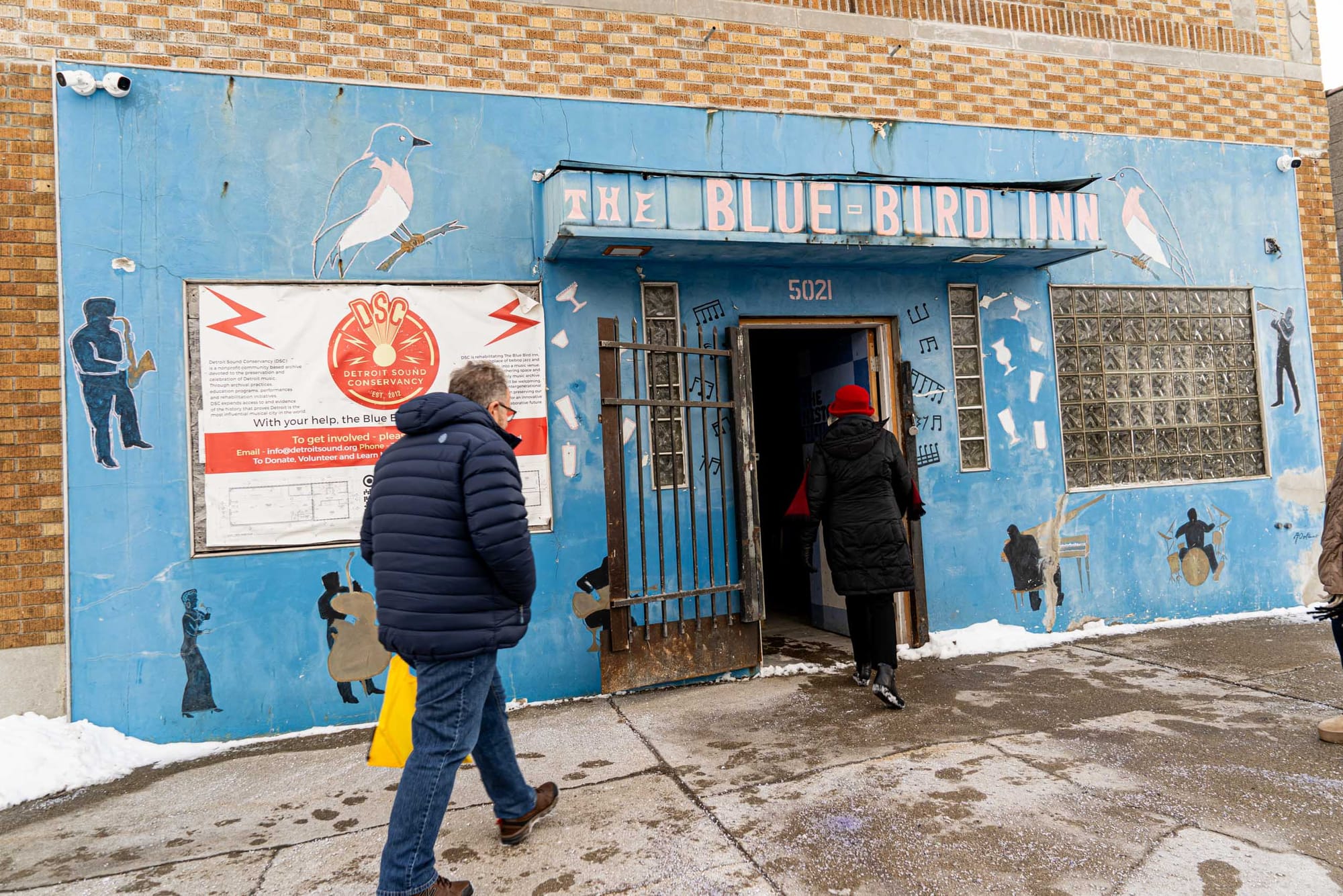 Two people in jackets walk towards a faded blue building with "The Blue Bird Inn" above the door.