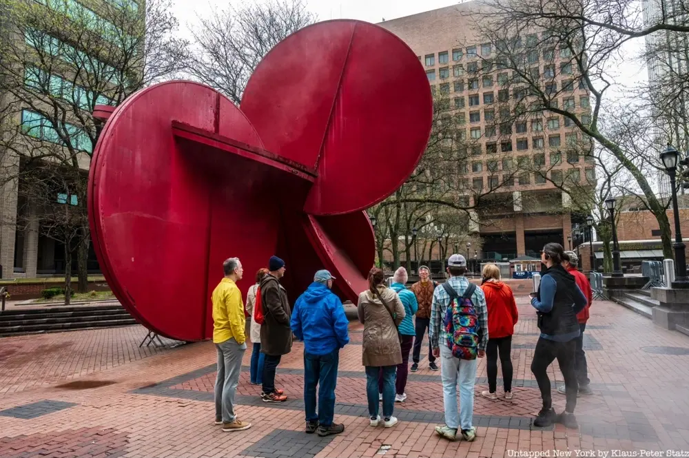 A group of people stand around a tour guide next to a large red circular sculpture.