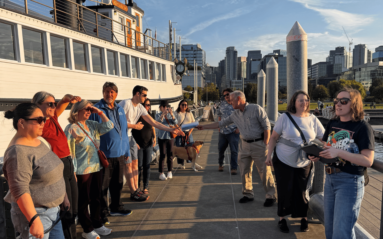 A dozen people stand listening on a pier with a big white boat behind them.