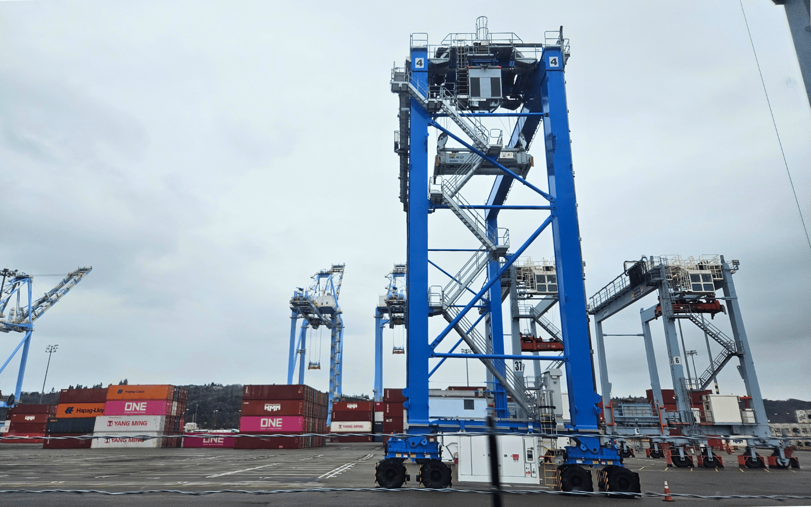 A tall blue machine with a staircase on the side in front of cranes and shipping containers.