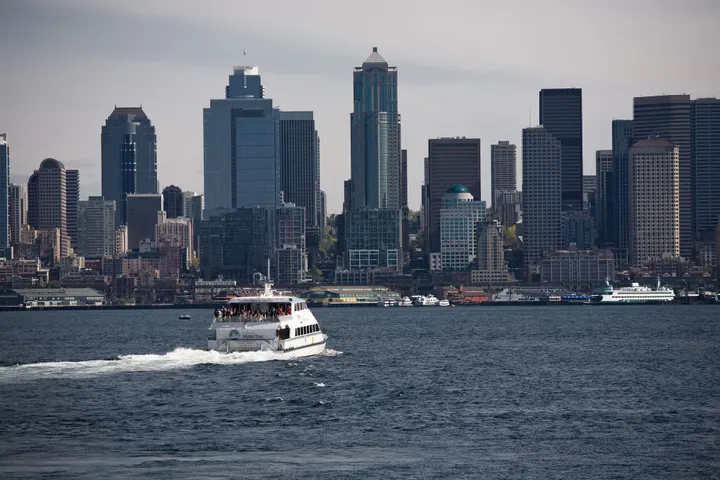A white vessel with passengers on the back deck crosses an open body of water towards a city skyline.
