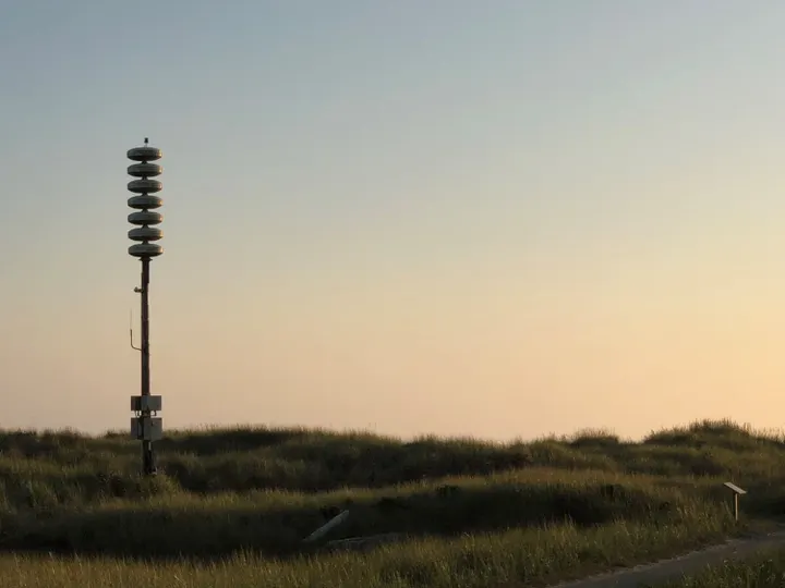 A pole with seven round devices stacked on top in a naturally grassy area.