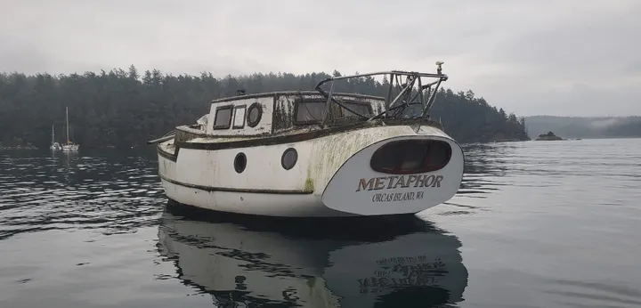 A weathered sailboat with no mast floats in front of a tree-covered island.