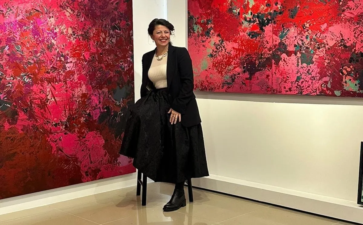 Artist Seçil Erel smiles on a stool between her large abstract paintings in a bright gallery. She wears a black blazer and skirt, flanked by a tall pink canvas and a wide red two-panel work.