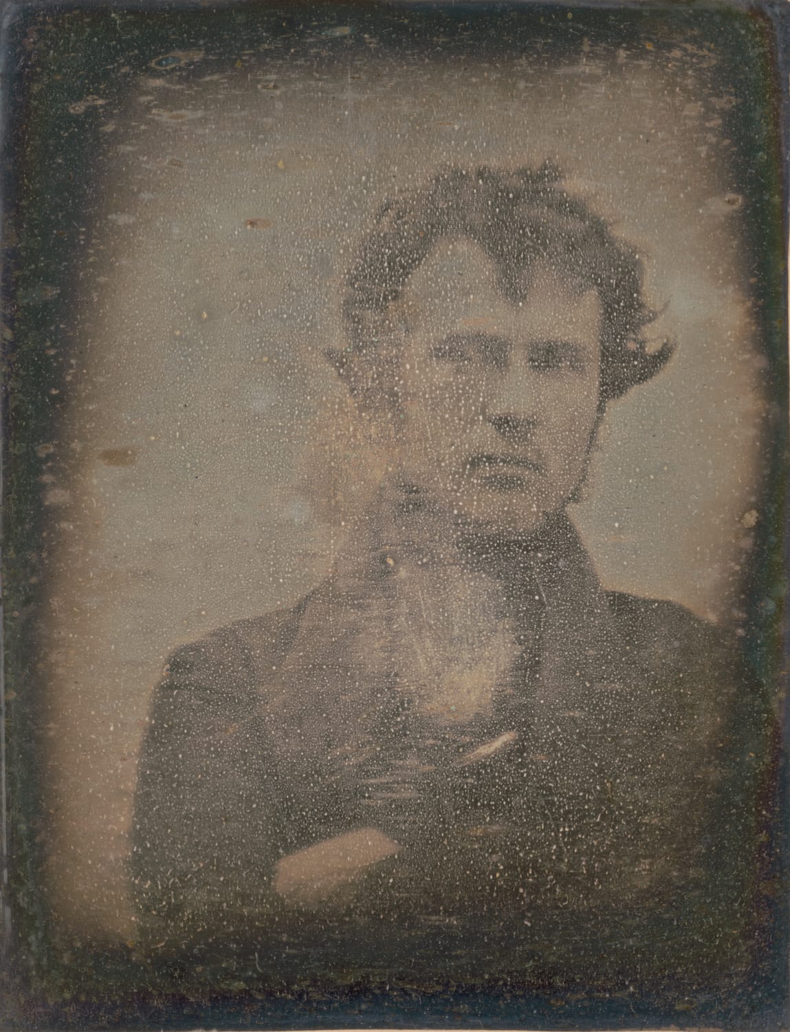 An old and spotted daguerrotype photograph of a young man, right hand tucked into the lapel of his jacket. He is looking slightly right and has tousled hair.