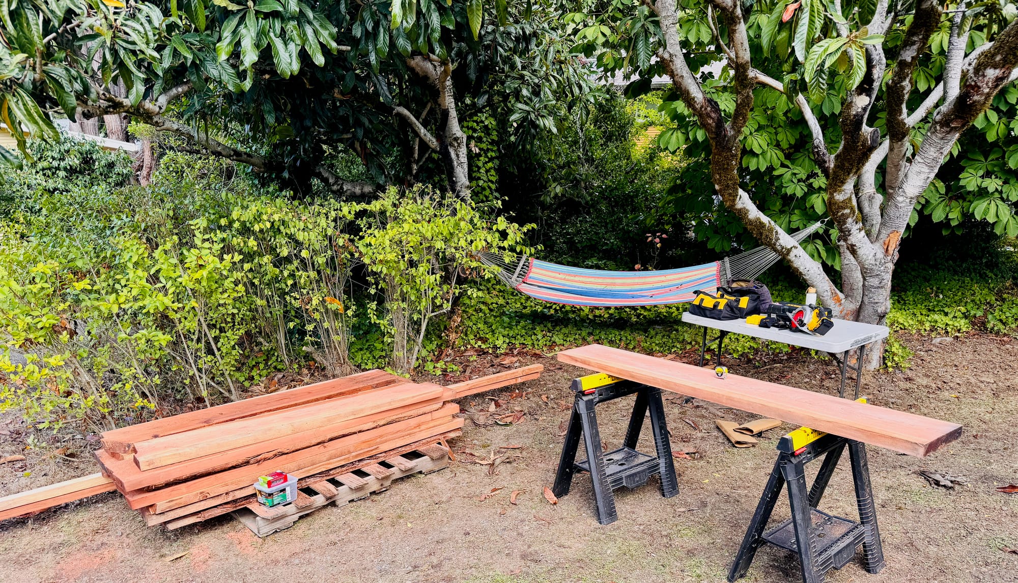 A backyard woodworking setup with a stack of reddish-brown lumber boards resting on pallets at left, and a single board laid across two black sawhorses at right. A folding worktable holds tools including what appears to be a sander and tool bag. A colorful striped hammock is strung between trees in the background, surrounded by lush green foliage.