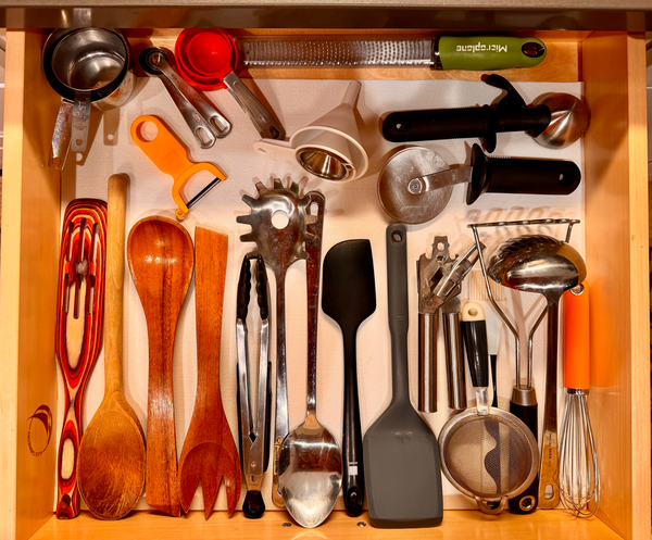 "A kitchen drawer filled with cooking utensils, laid out flat and organized by size and type on a white liner."