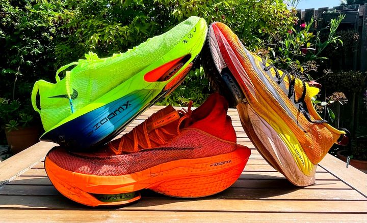 Three pairs of Nike ZoomX Alphafly running shoes in bright neon colours stacked on a wooden table outdoors — popular carbon-plated racing shoes among sub-3 marathon runners