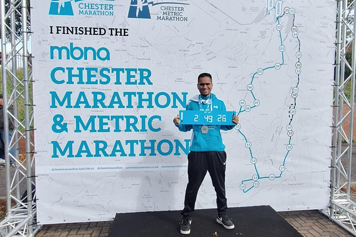 George Fernandez stands proudly at the Chester Marathon finish area holding a sign showing his time of 2:49:26, wearing his medal and blue finisher’s hoodie.