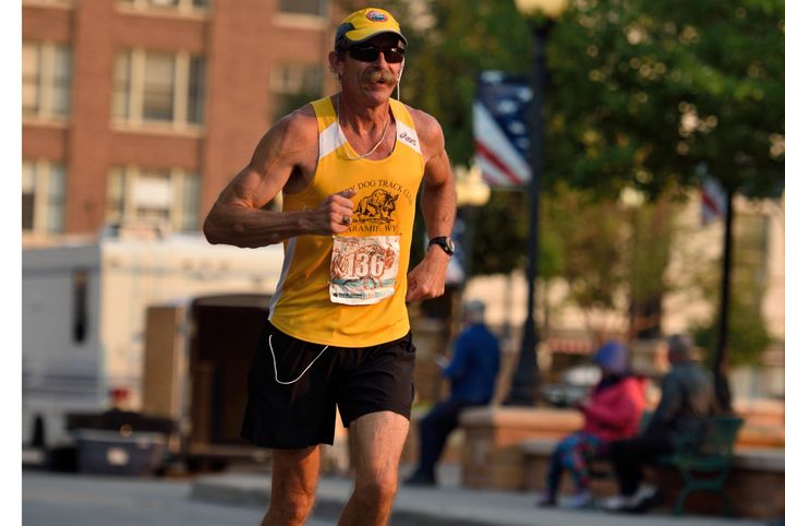 Jerry Rief running a road race in a yellow Laramie, Wyoming track-club vest and cap, mid-stride on a city street with spectators and buildings blurred in the background.