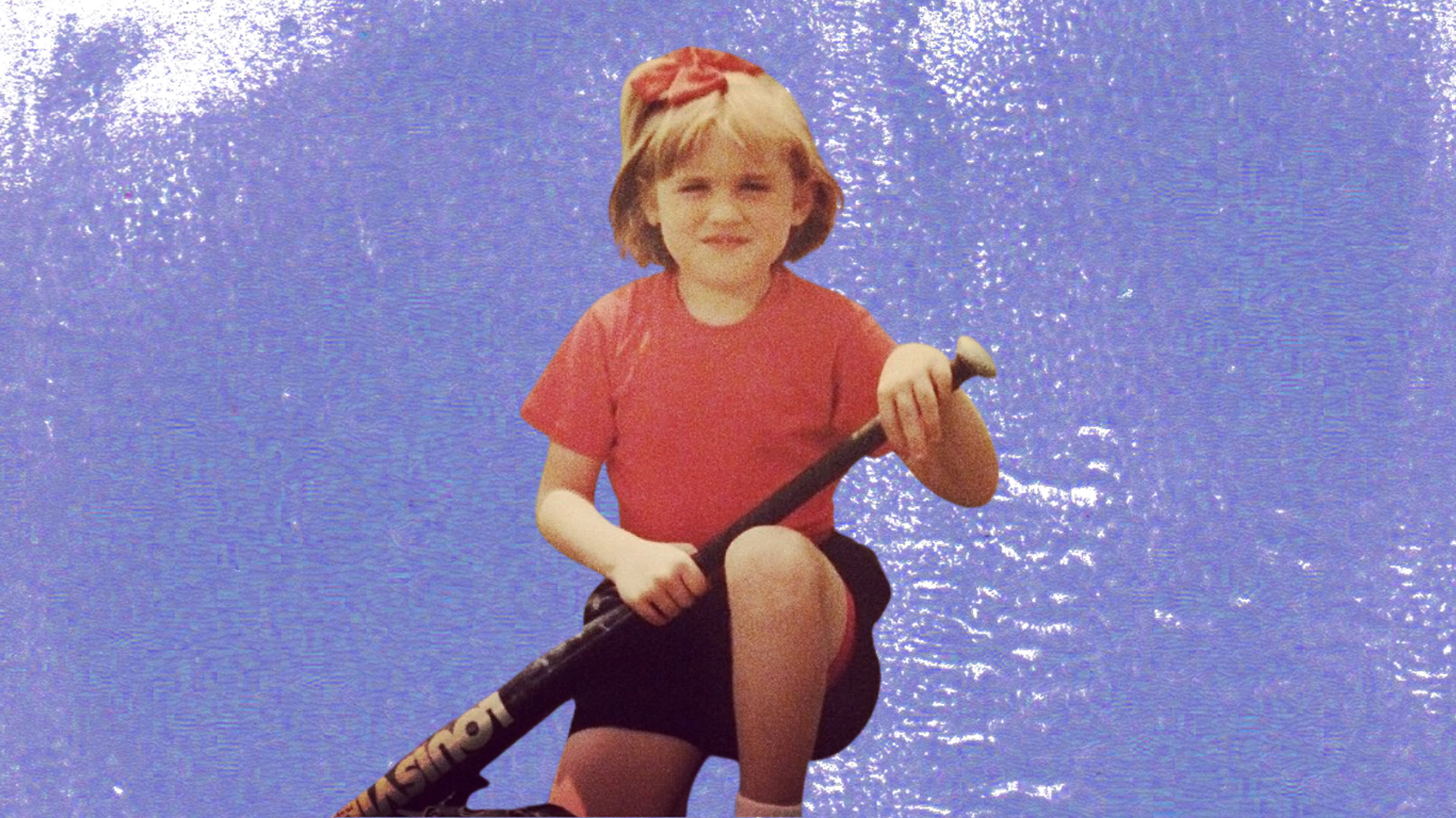 Amy McCarthy as a child, in a red tshirt, black shorts and a red bow, holding a baseball bat in front of a purple background