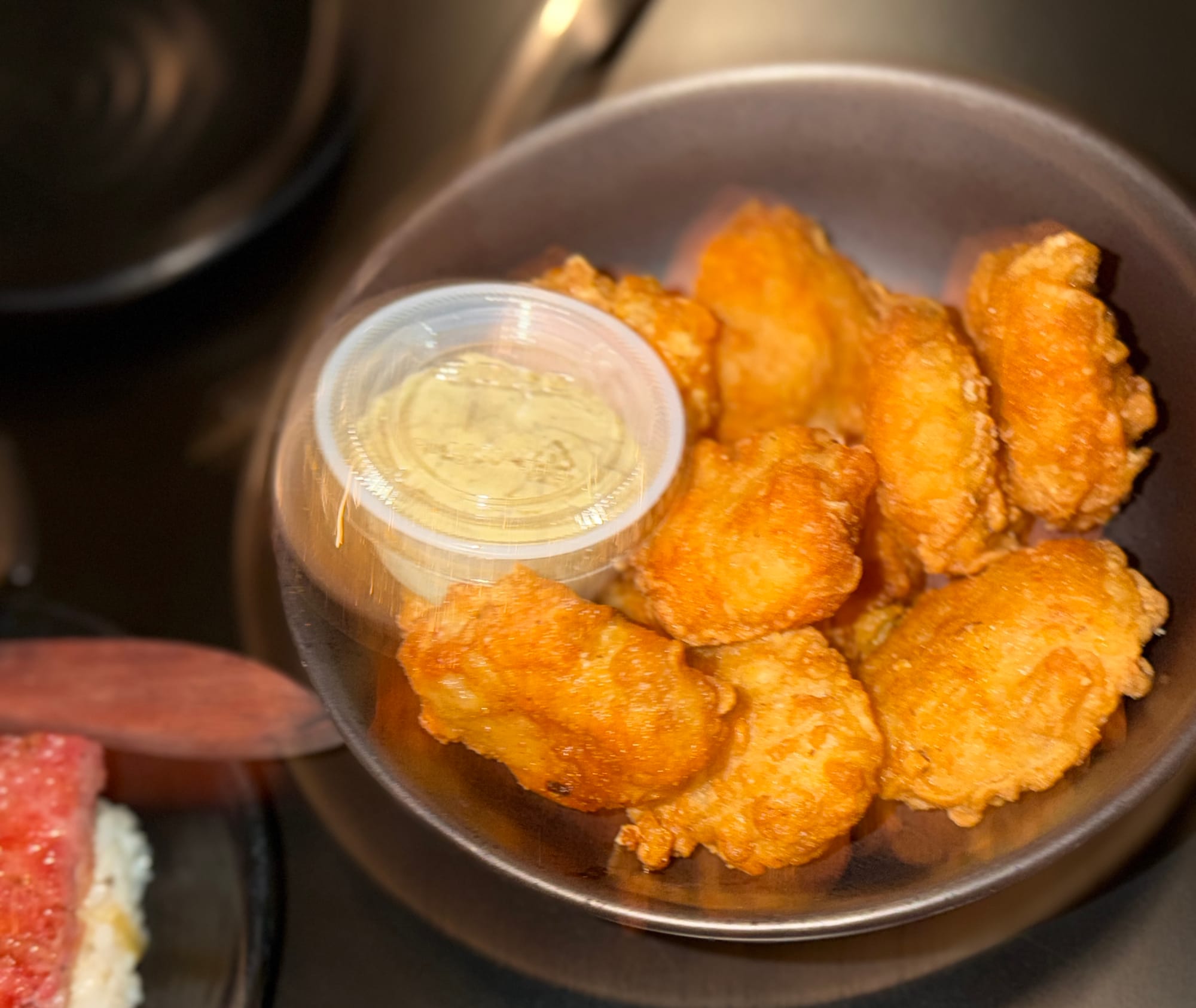 A black bowl holds fried chicken nuggets and a plastic ramekin of sauce