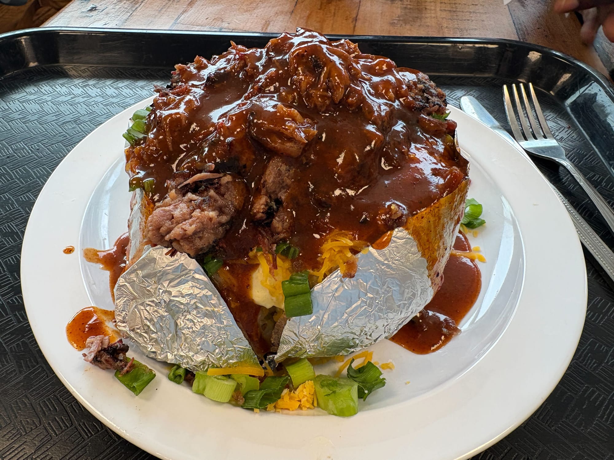 a baked potato wrapped in tinfoil and topped with brisket, cheese and scallions, on a white plate