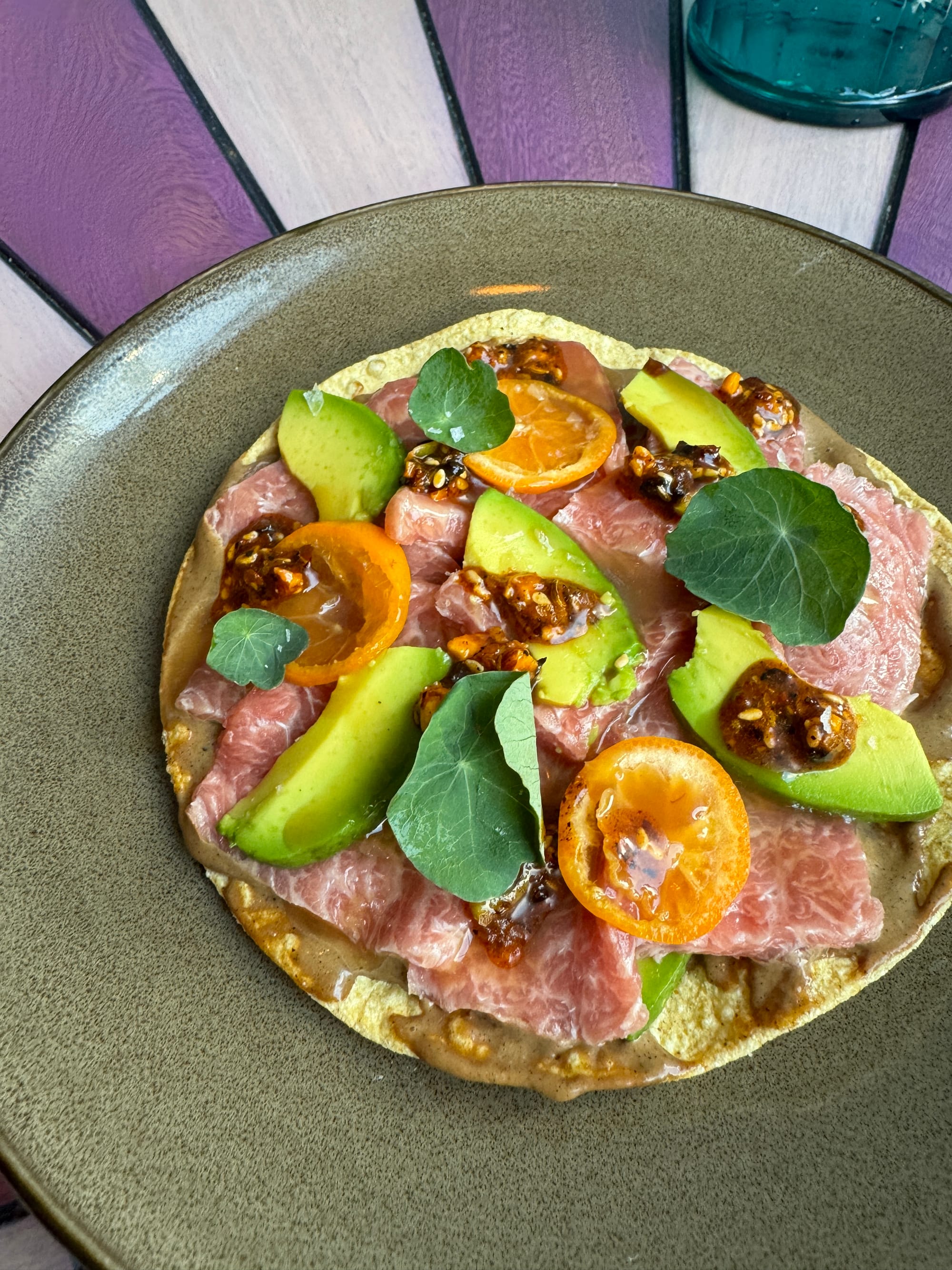 A tuna tostada topped with slabs of tuna, avocado slices, and small golden tomatoes.