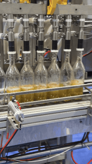 Six bottles being filled with a yellow liquid by a factory machine.