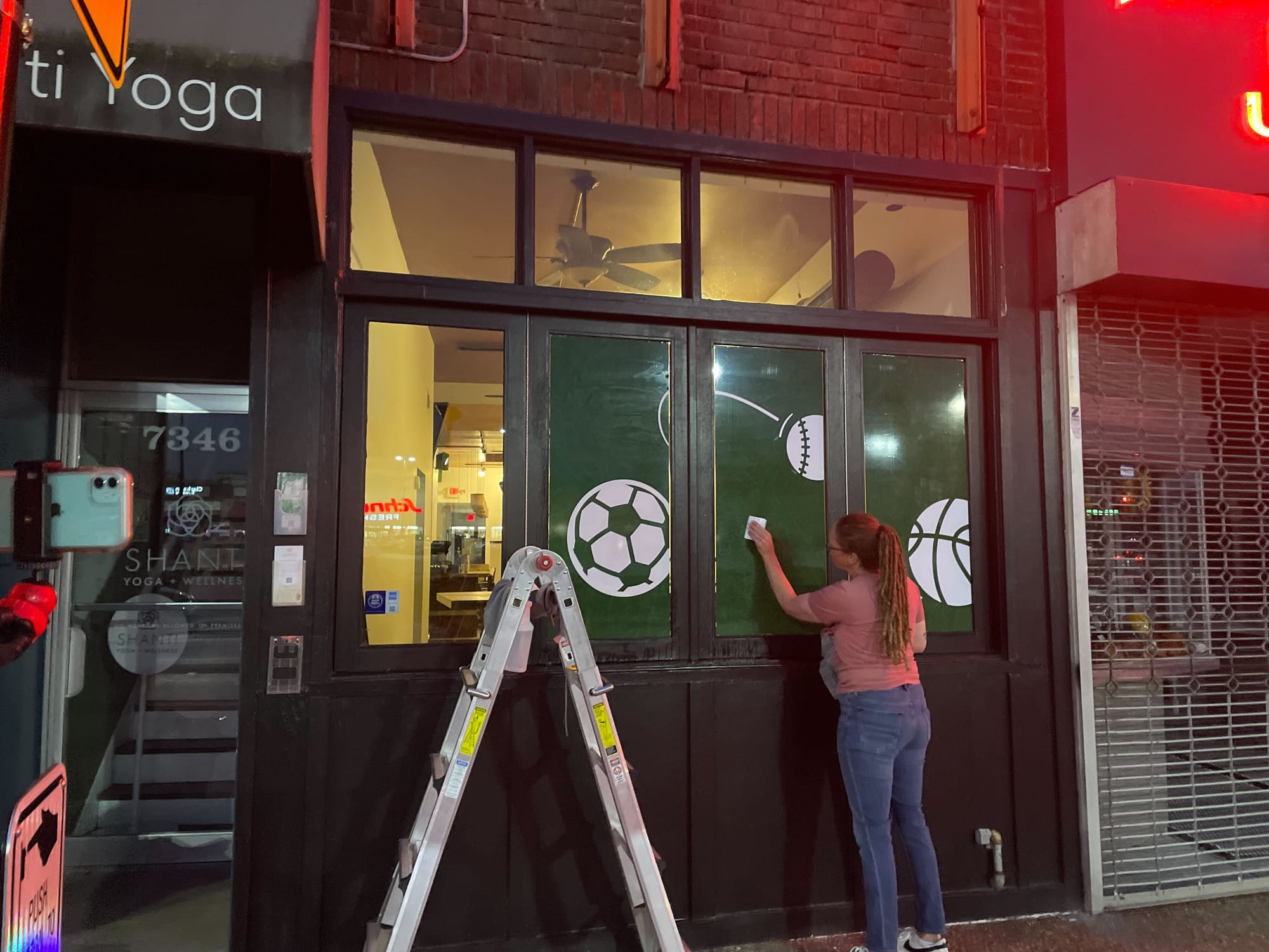 A woman, at night, putting window clings onto a bar's exterior window with a ladder on the sidewalk.