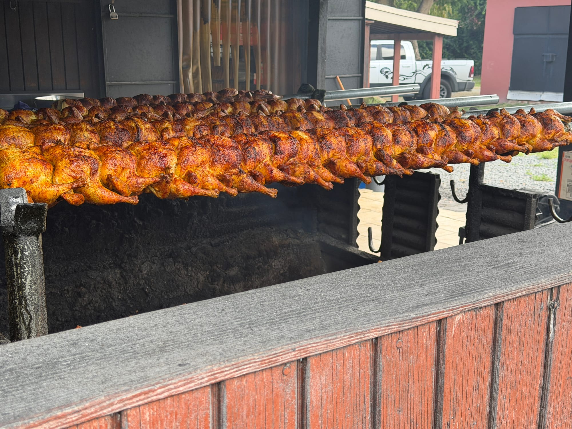 Multiple rows of rotisserie chickens hang over an open pit