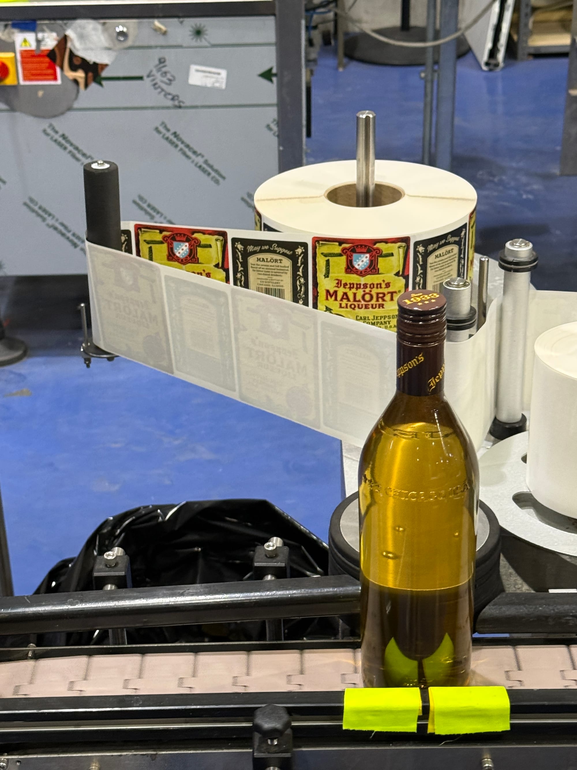 A brown glass bottle on a conveyor belt, next to a labeler of Malört labels