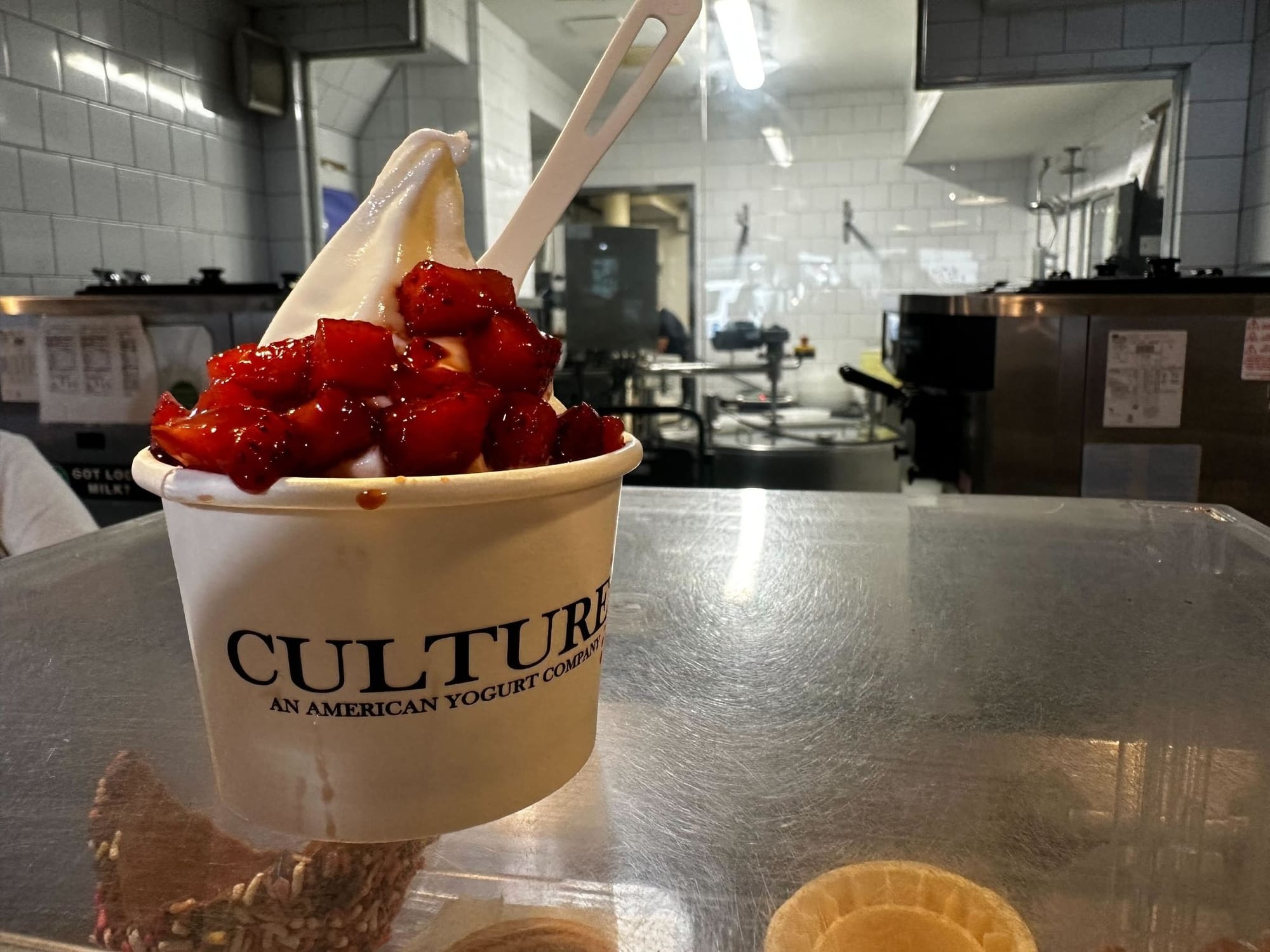 A paper cup full of white frozen yogurt with strawberries on a steel counter.