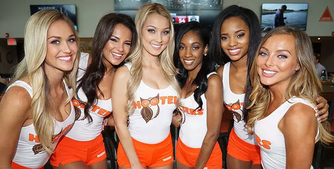 Five women wearing white tank tops that say Hooters, and orange shorts