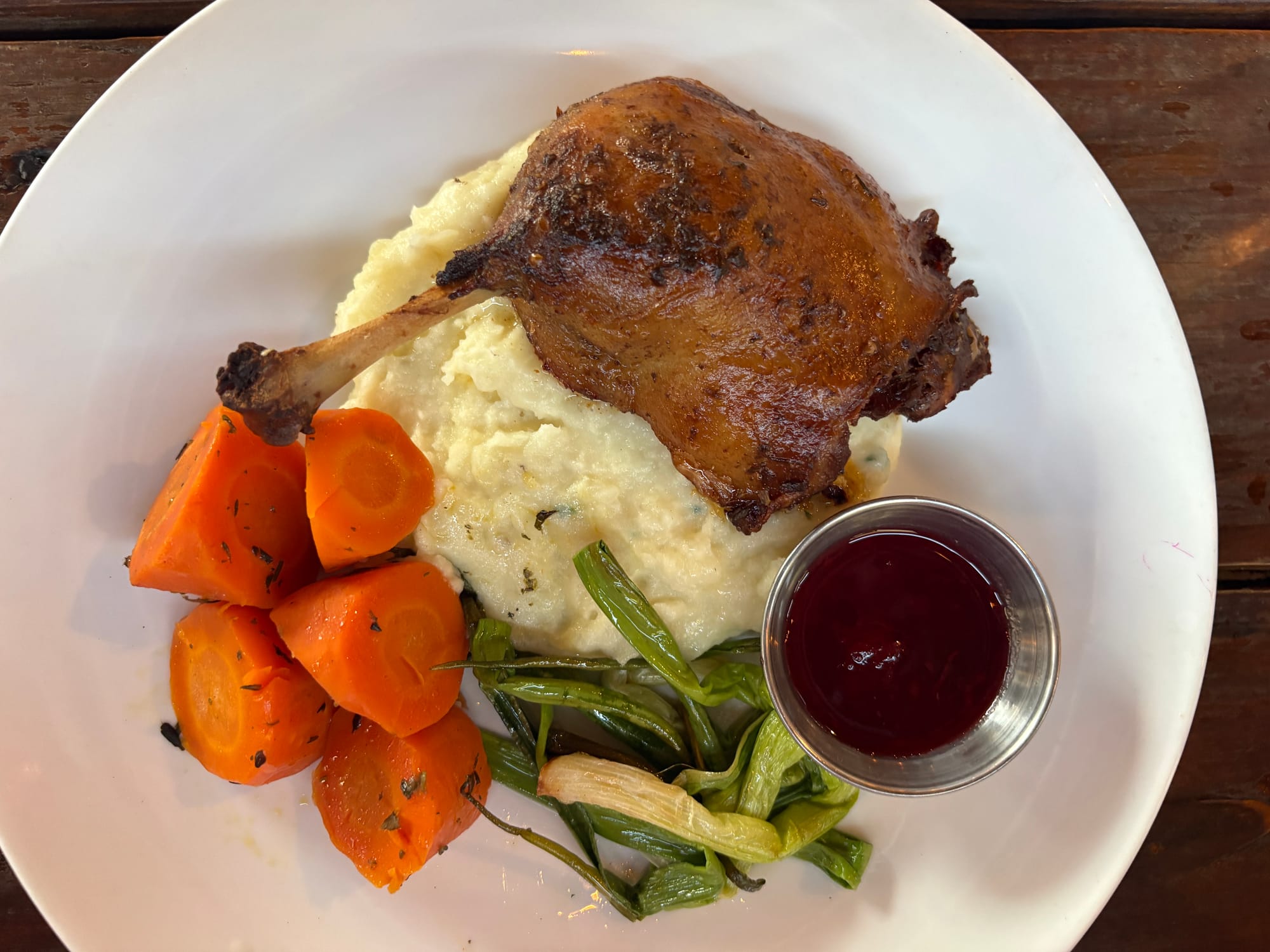 A plate holds a duck leg, mashed potatoes, sauce, green onions, and carrots.