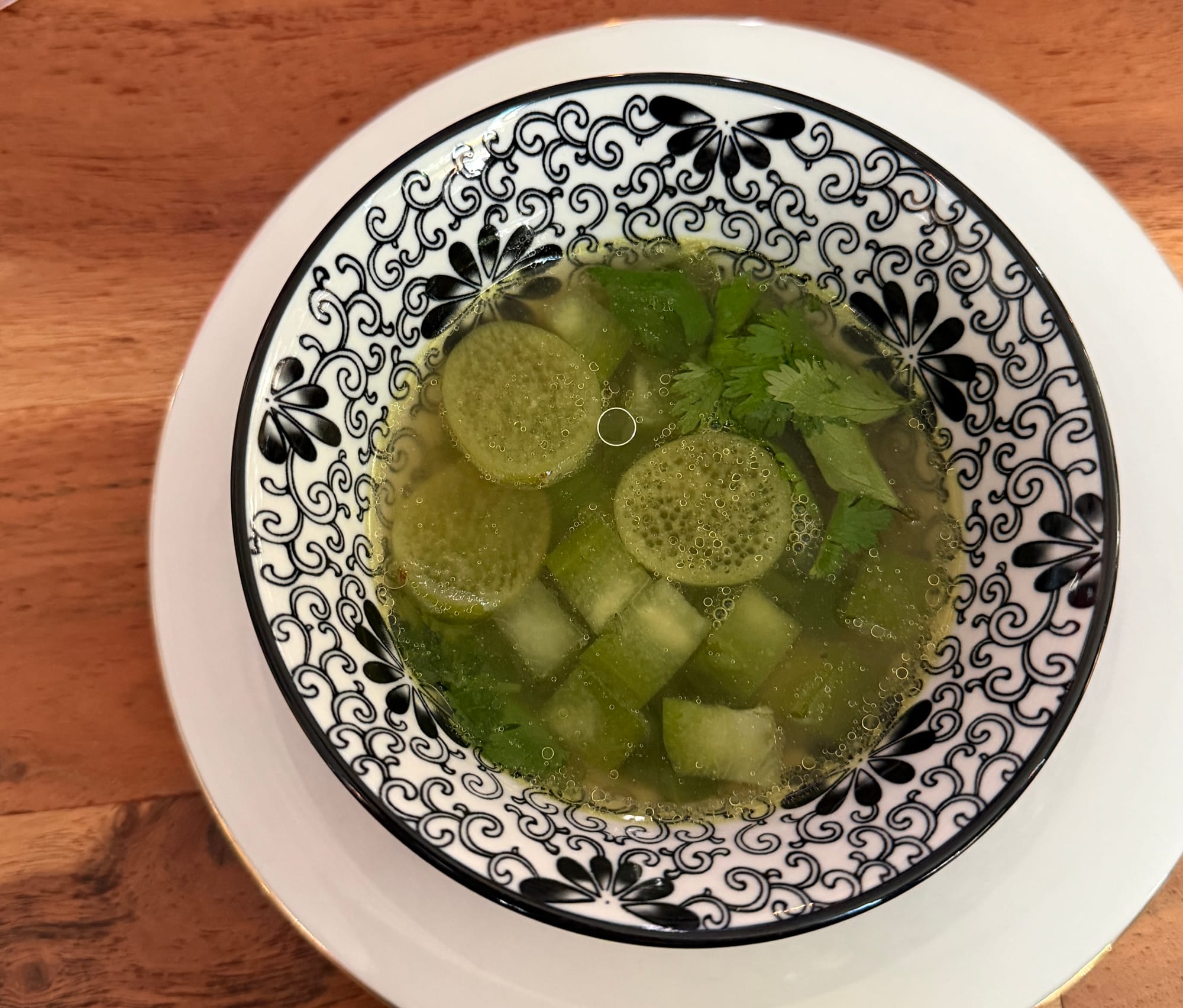 A blue and white patterned bowl of broth, herbs, and green vegetables sits atop a wood table.
