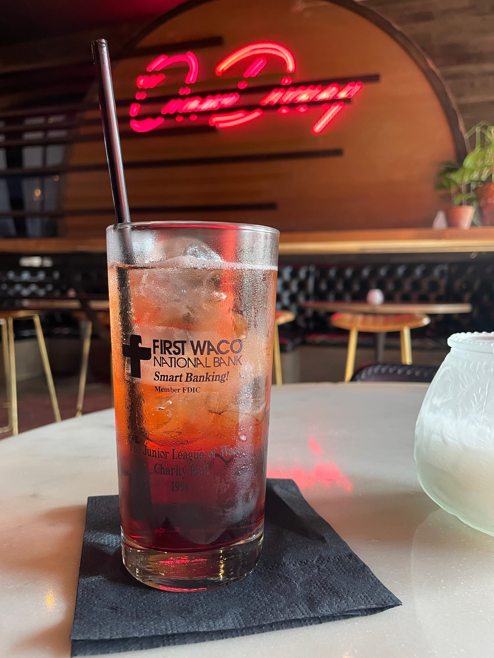 A glass with a brownish cocktail sits in front of a neon sign in a bar that reads One Day.
