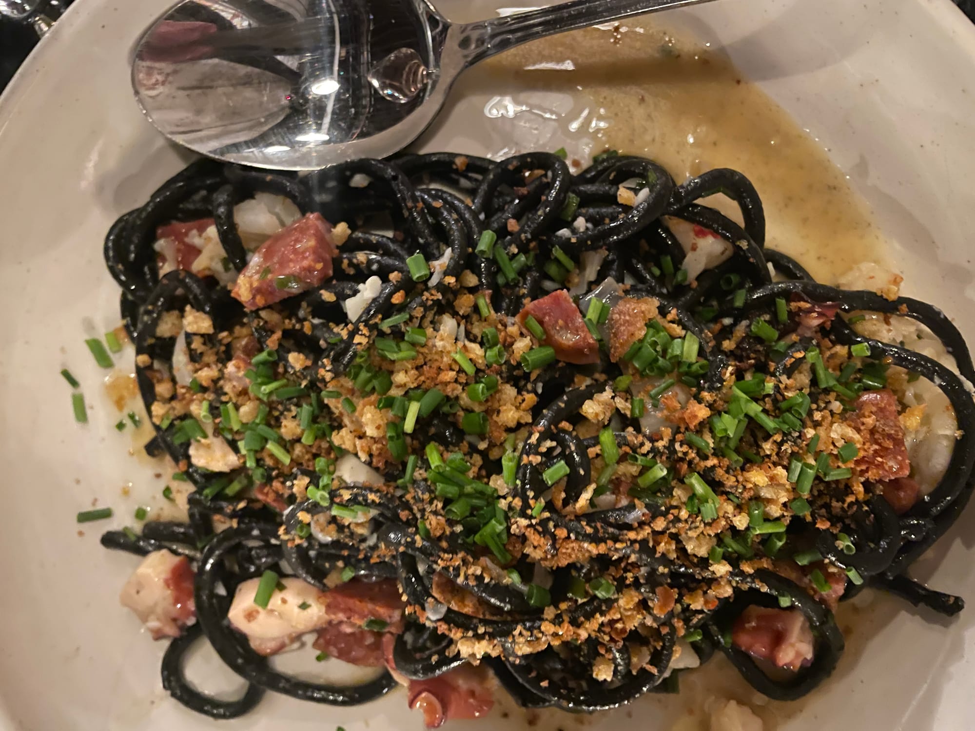 A plate holds squid ink pasta in a burre sauce with chives, bread crumbs, and chunks of pork.