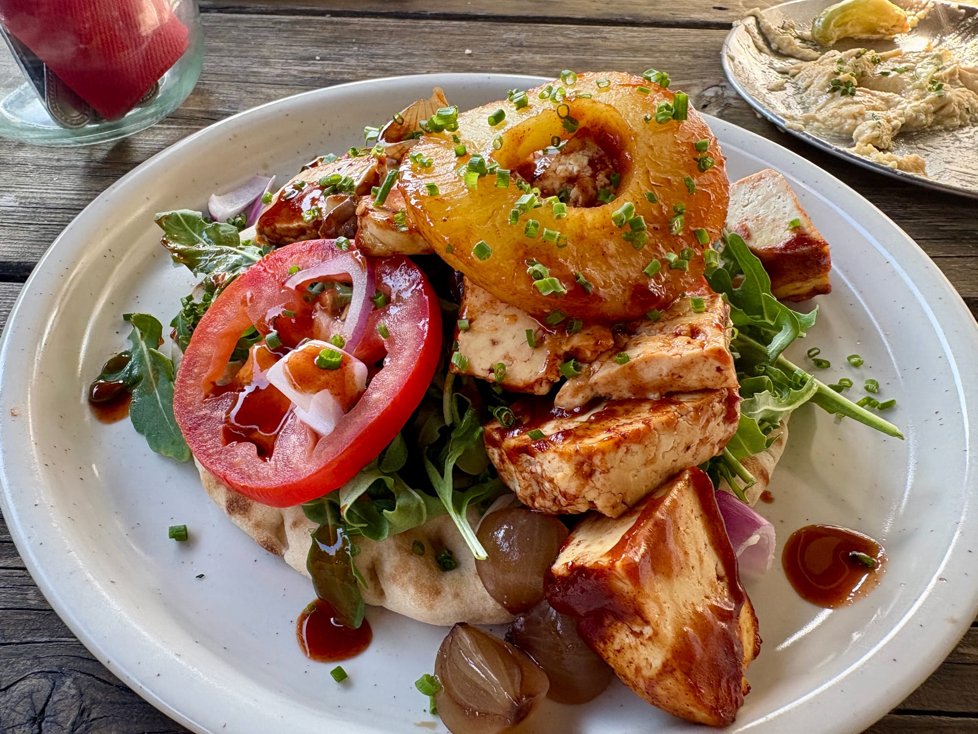 A plate holds a ofu and pineapple flatbread loaded with arugula, tomatoes, and pearl onions.