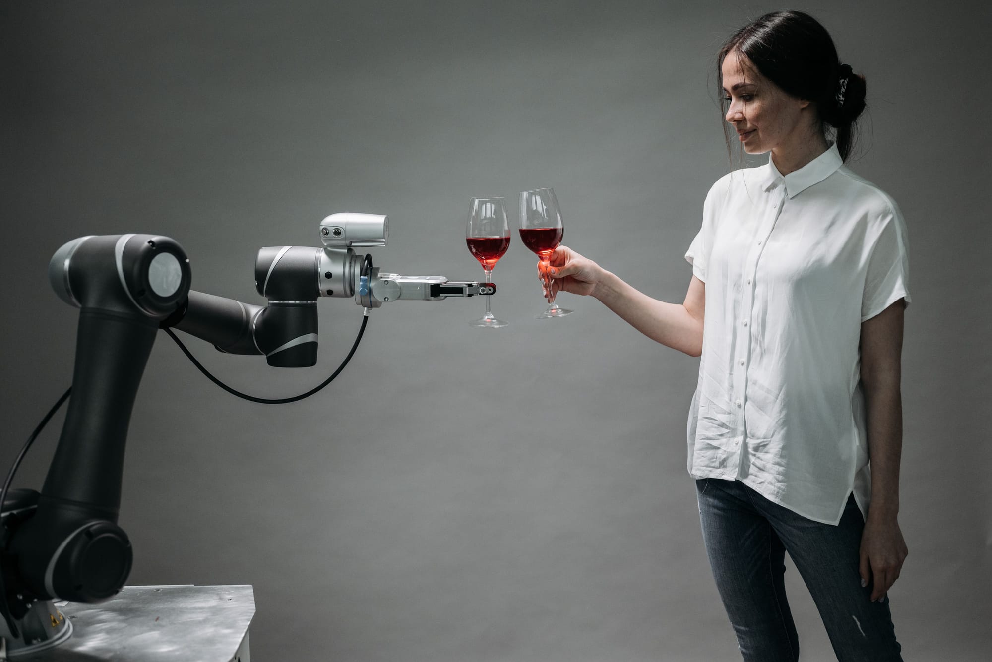 A woman in a white button down shirt and jeans cheerses glasses of wines with a robotic arm.