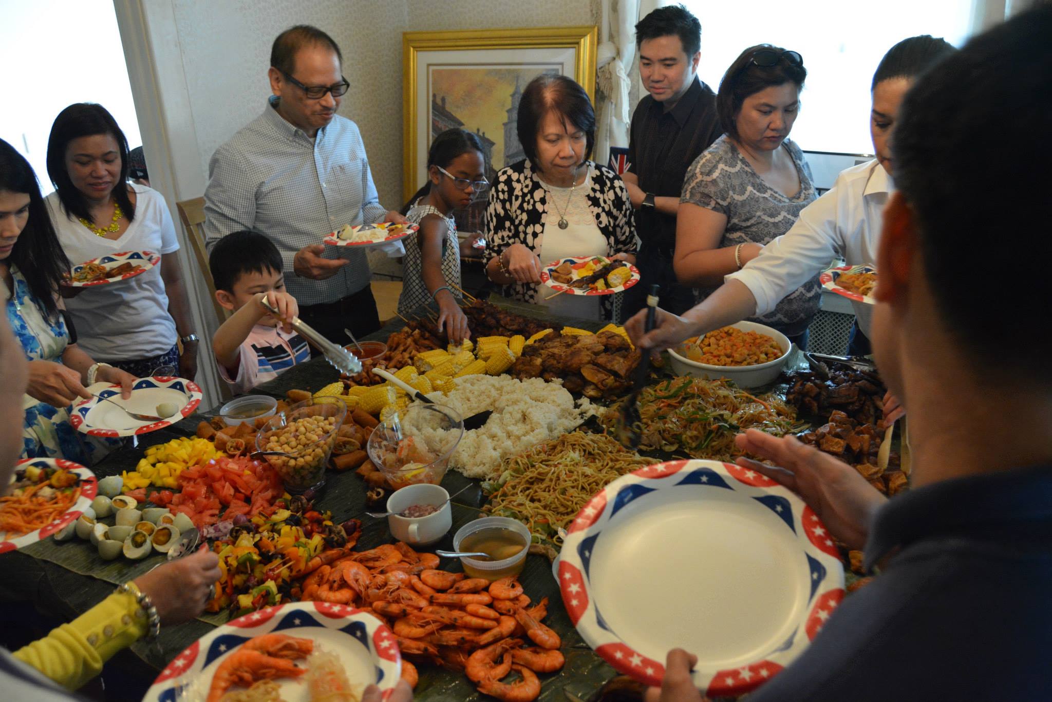 A photo showing a kamayan feast with various Filipino foods sitting on banana leaves. The food is surrounded by Filipinos grabbing food for their plate.
