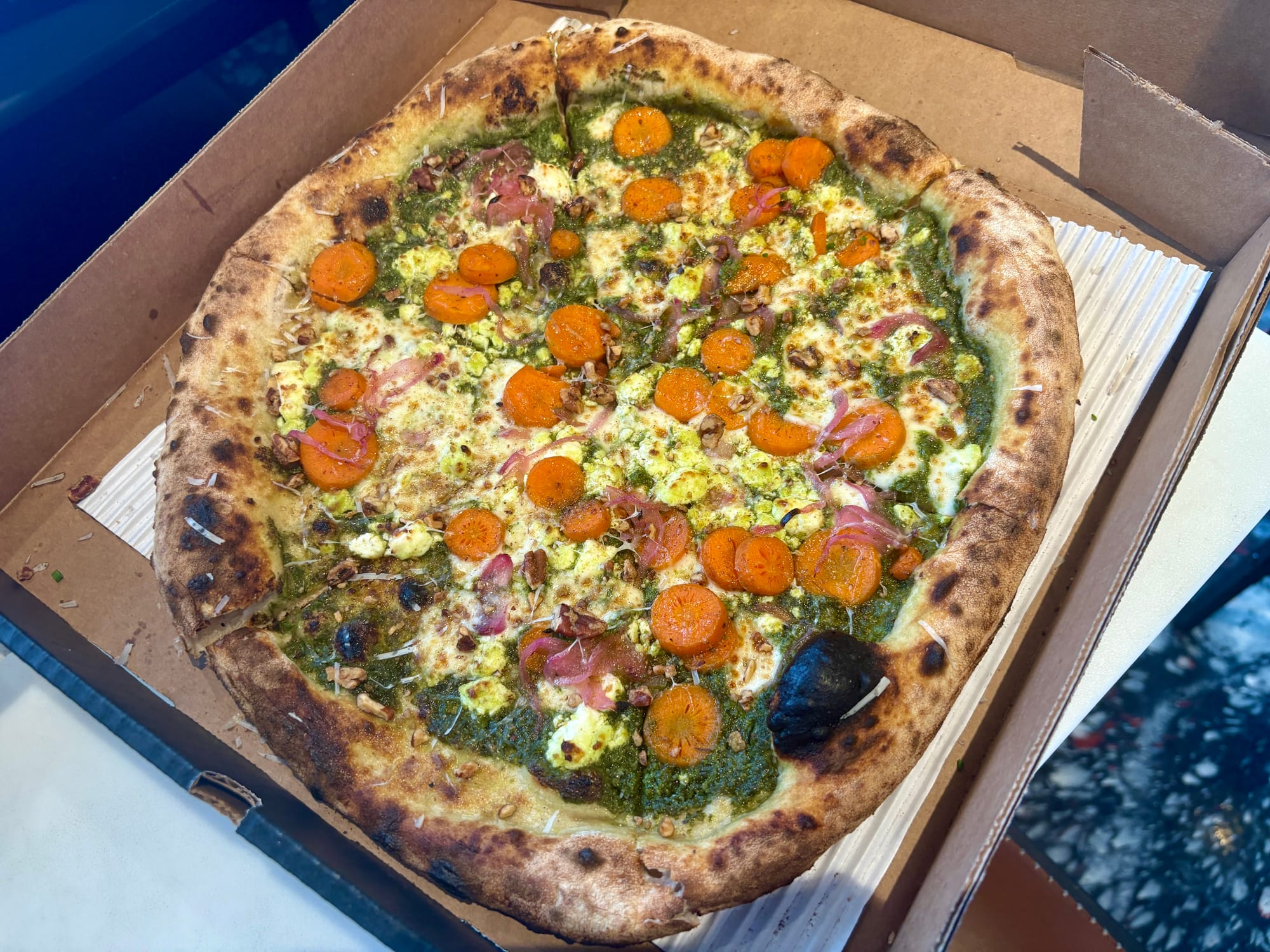 A box holds a pizza topped with carrots, red onion, and basil sauce.