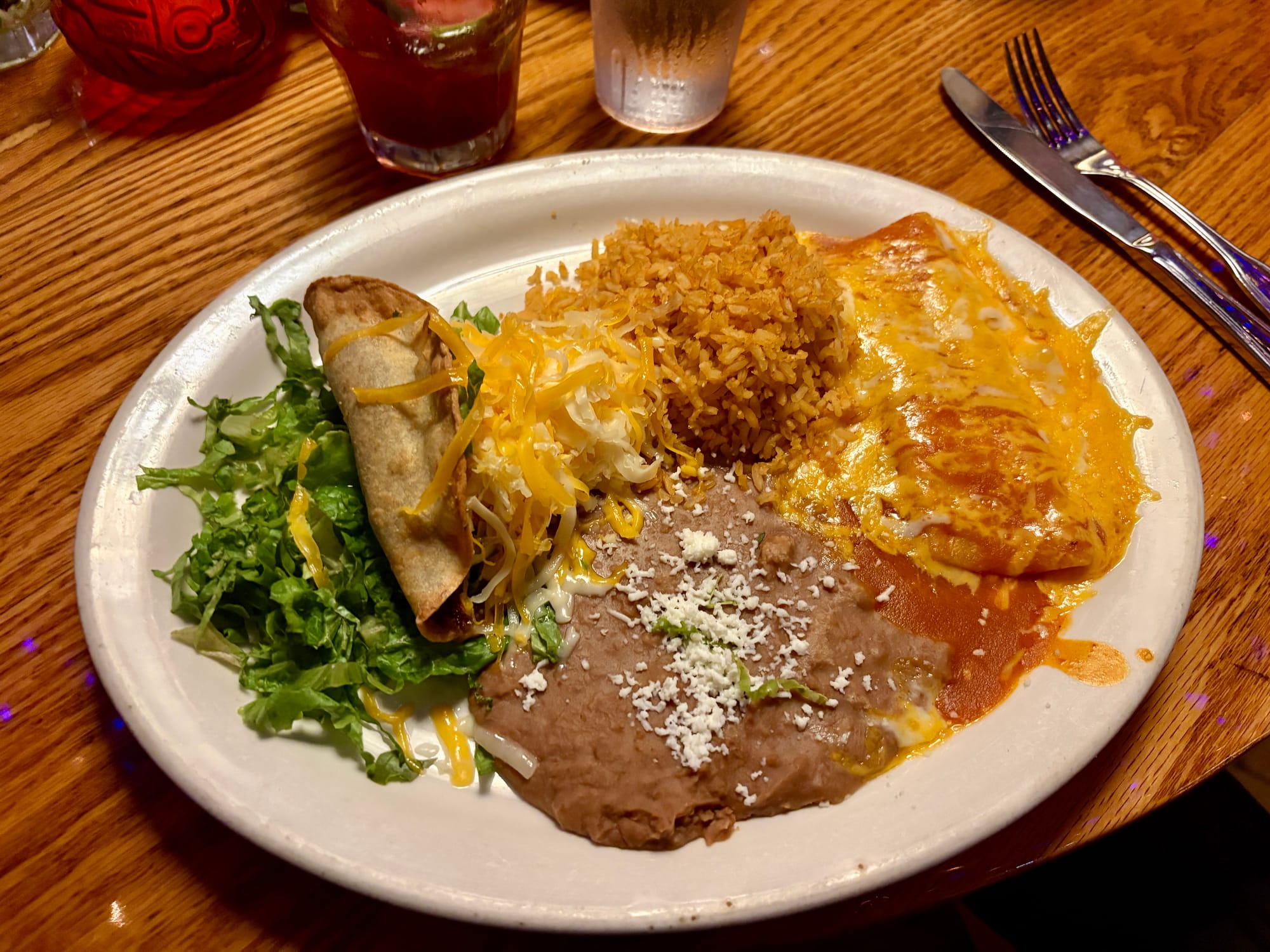 A plate of Mexican food holds, left to right, a pile of shredded lettuce, a rolled taco with lots of shredded cheese, Spanish rice on the top, refried beans on the bottom, and one cheese enchilada with red sauce.