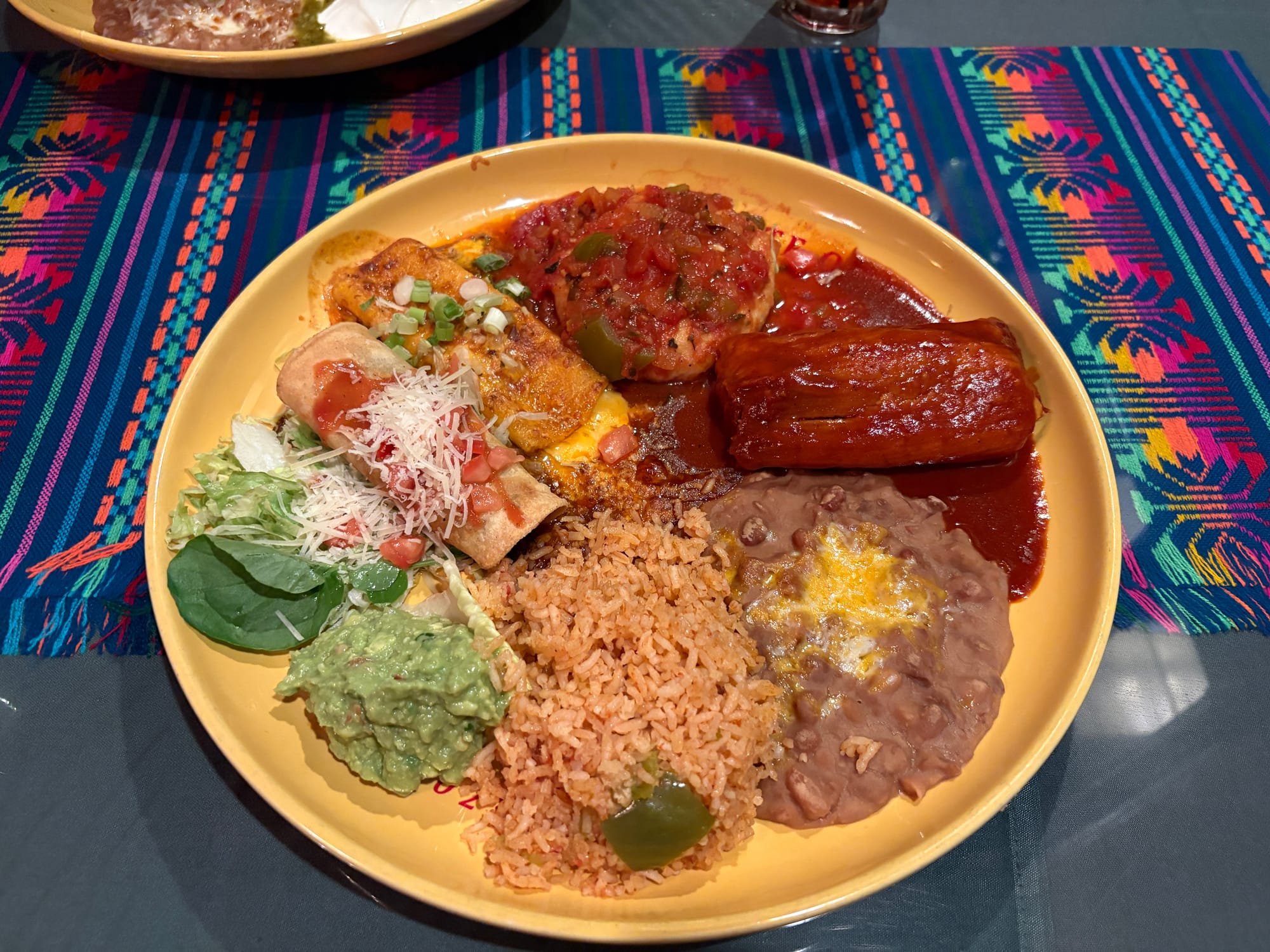 A circular plate of Mexican food holds, clockwise from top, chile relleno, a beef tamale in dark red sauce, refried beans with cheese, Spanish rice, guacamole, a rolled taco, and a cheese enchilada.