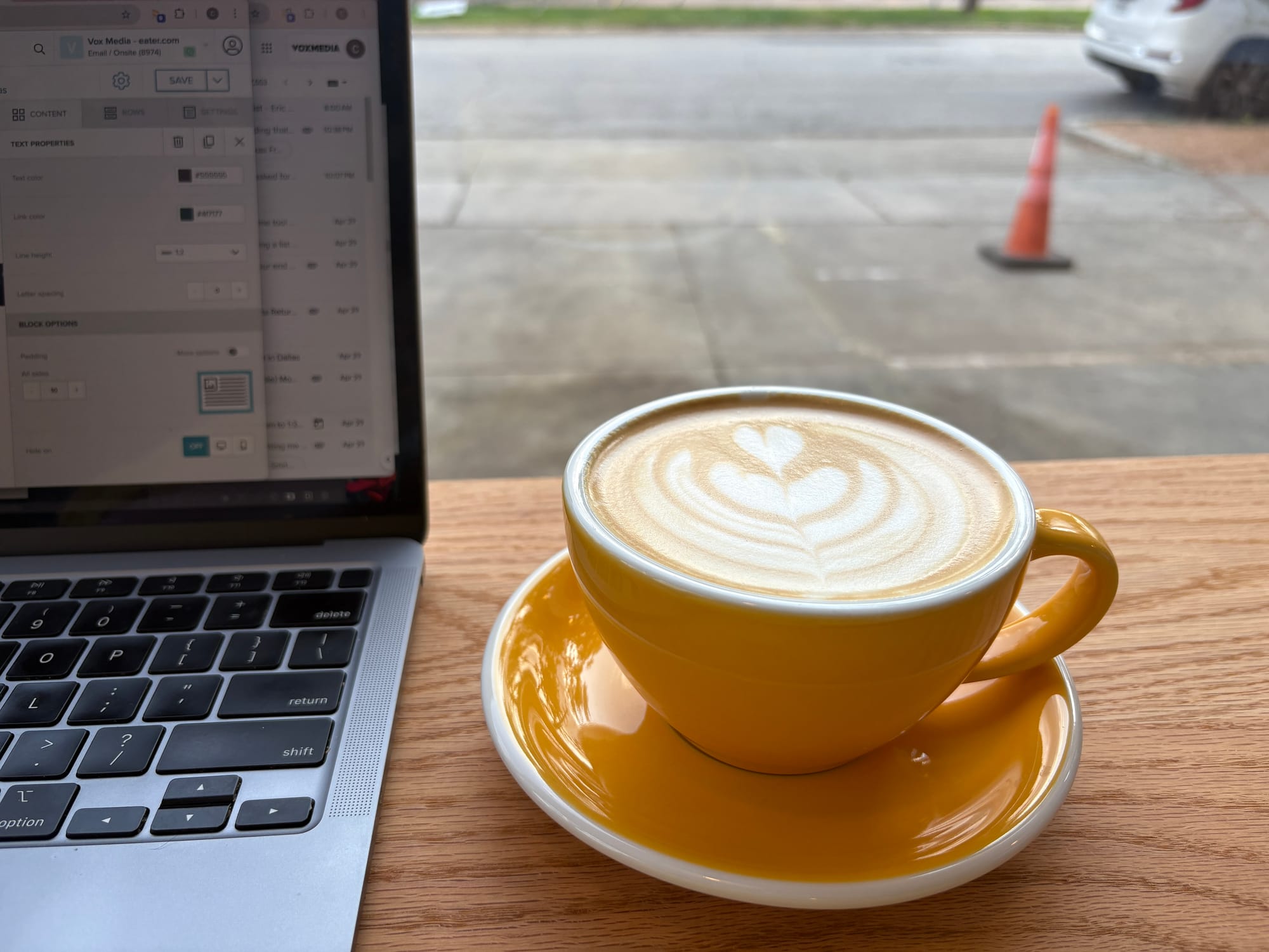 An orange cup of coffee with hearts in the milk sits next to an open laptop looking onto a parking lot.