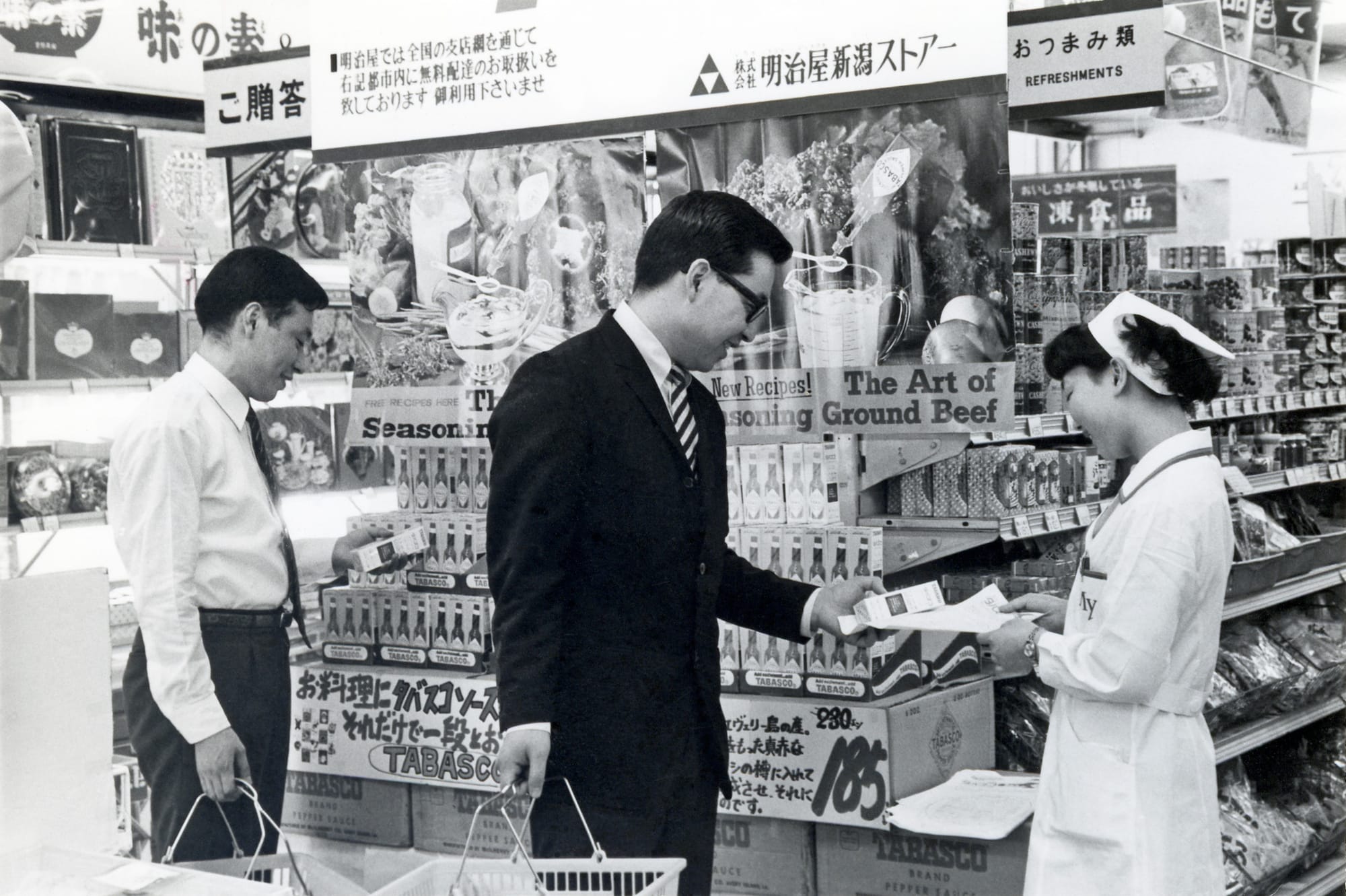 A black and white image of two men and a woman standing in front of a Tabasco display in a Japanese grocery store in 1970.