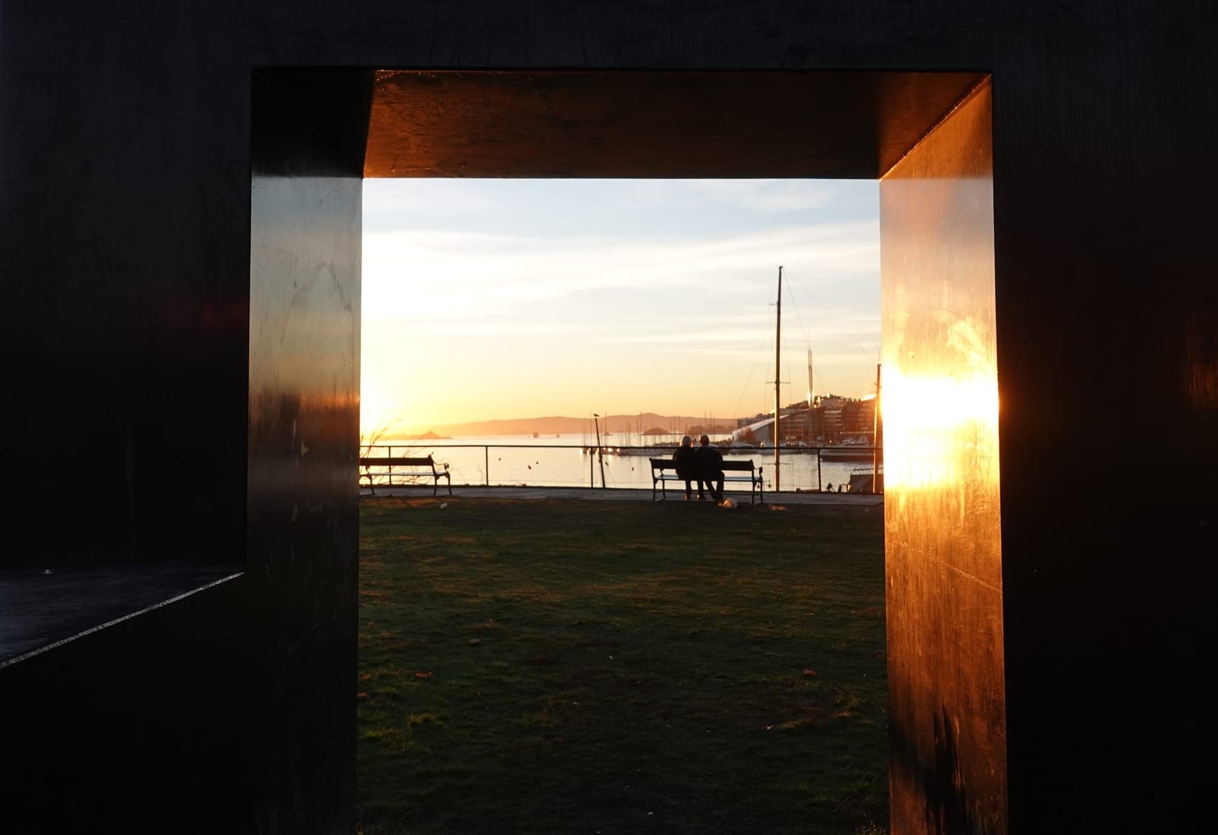 A photo through a monument of people on a bench overlooking the harbour, Oslo, Norway