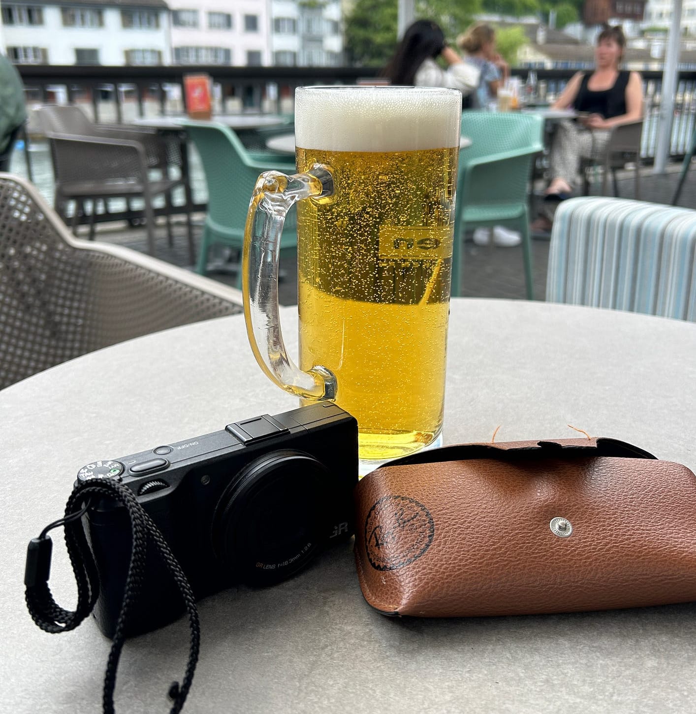 A photograph of a pint of beer on a table next to a camera and sunglasses