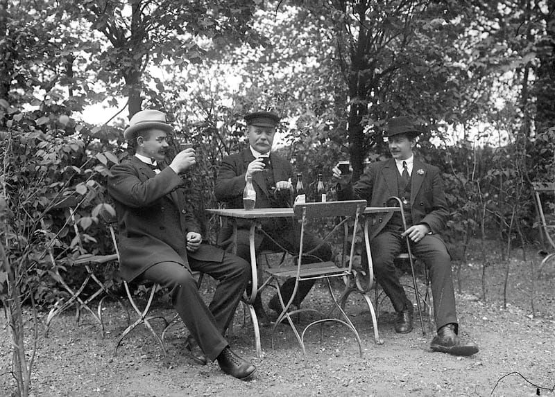 An old photo of three men sat drinking beer - Tre män som sitter utomhus vid ett bord och dricker öl och sockerdricka, vy 1920 på asken 	Metallutfällning. by Johnson Julius fotograf - Trelleb