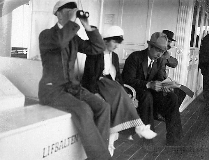 A photo of a man on a boat looking through binoculars - Enligt noteringar: "Juni -19." by Okänd - Bohuslän Museum, Sweden - Public Domain.