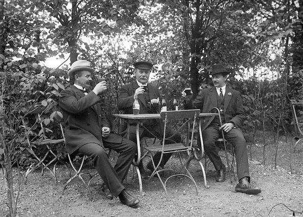 An old photo of three men sat drinking beer - Tre män som sitter utomhus vid ett bord och dricker öl och sockerdricka, vy 1920 på asken 	Metallutfällning. by Johnson Julius fotograf - Trelleb