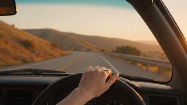 A photo taken from inside a car, looking through the windshield at a California road curving ahead, with one hand on the steering wheel.