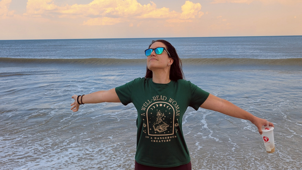 Dark haired woman with light skin, arms open to the sky, with the ocean in the background.