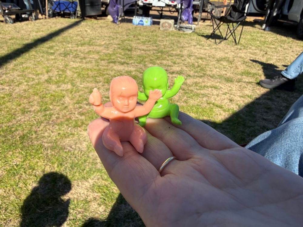 Two plastic king cake babies in a woman's hand. The closer baby is flesh-toned, the one in the background is green.